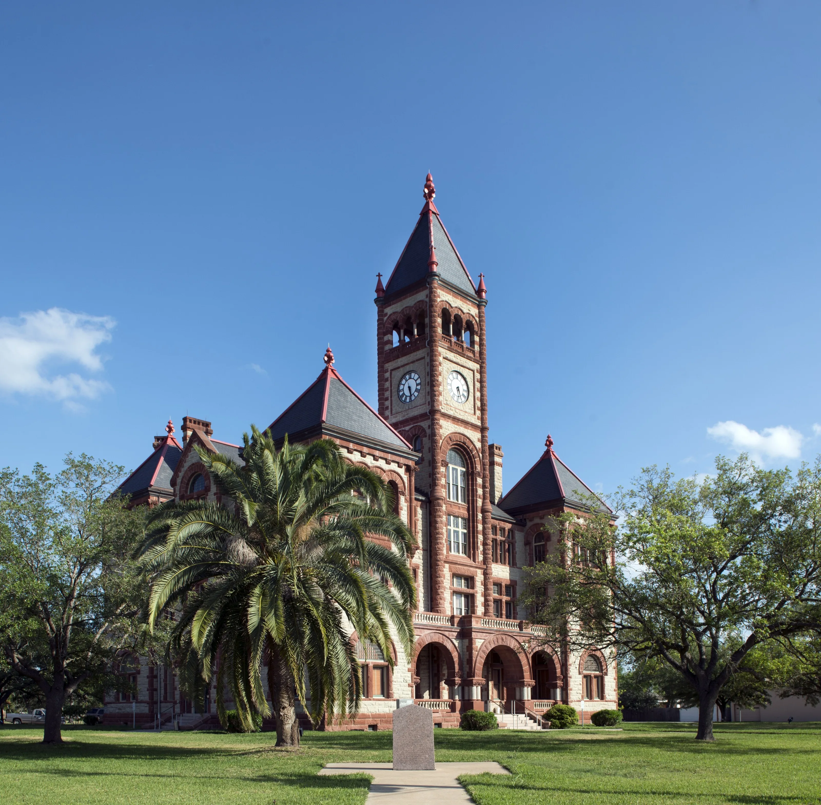 The red-brick DeWitt County Courthouse in Cuero, with a tall clock tower and turrets, behind a palm tree and lawn under a clear blue sky.