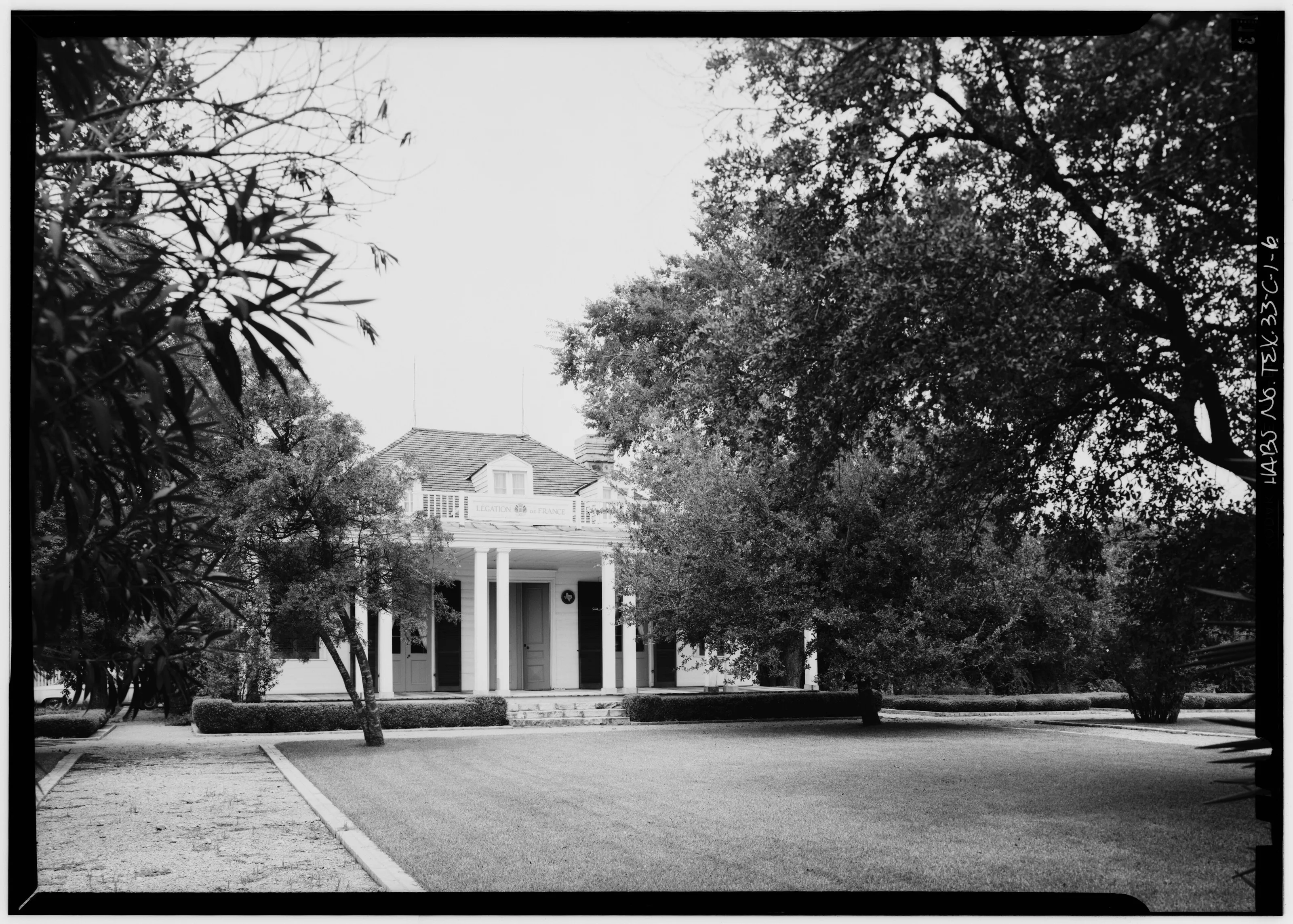 Black-and-white photograph of the French Legation house in Austin, a white frame building with a columned porch, set back behind a drive and large trees.