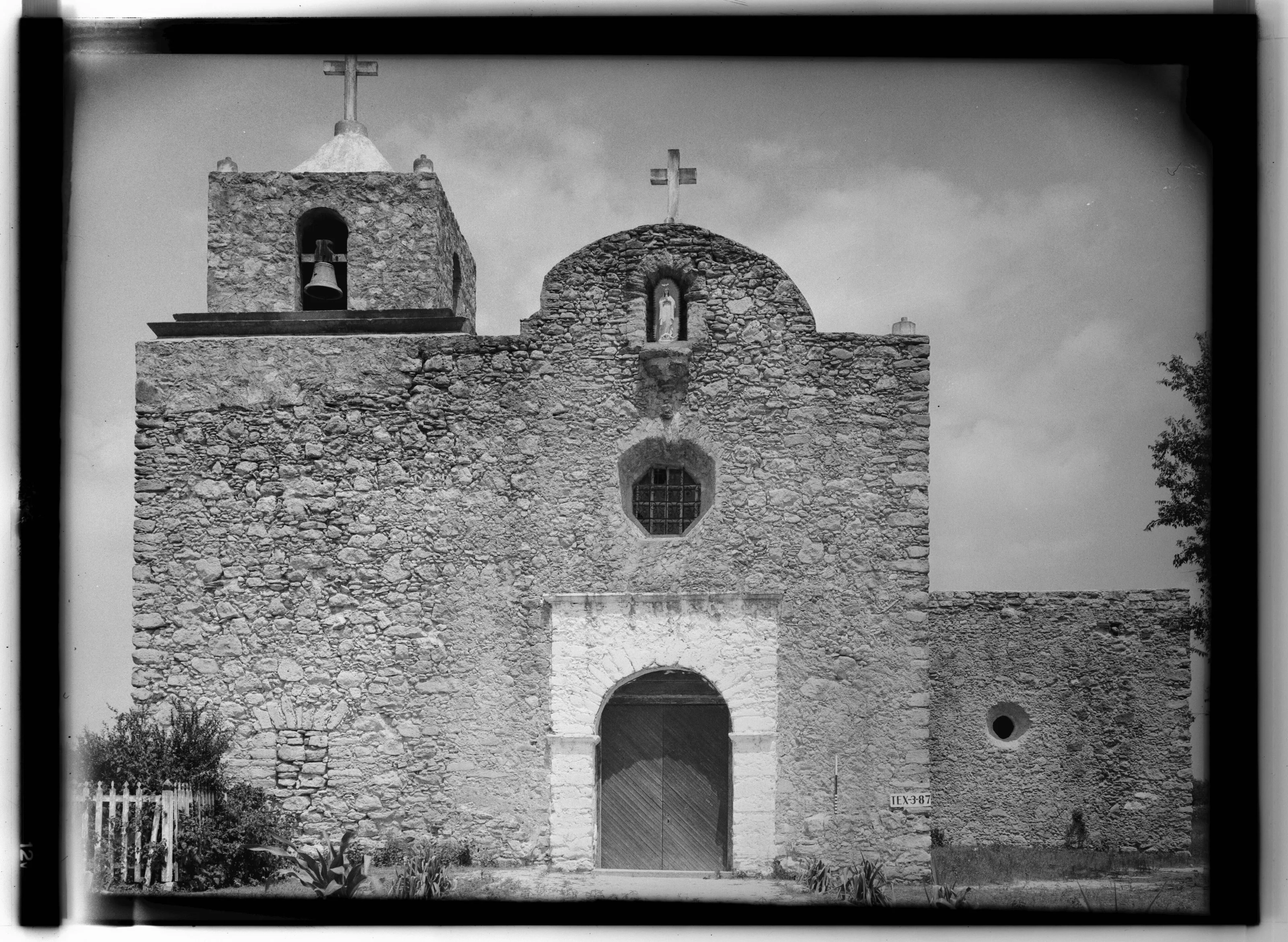 Black-and-white photograph of the stone chapel at Presidio La Bahia with its bell tower, round window, and arched wooden door.