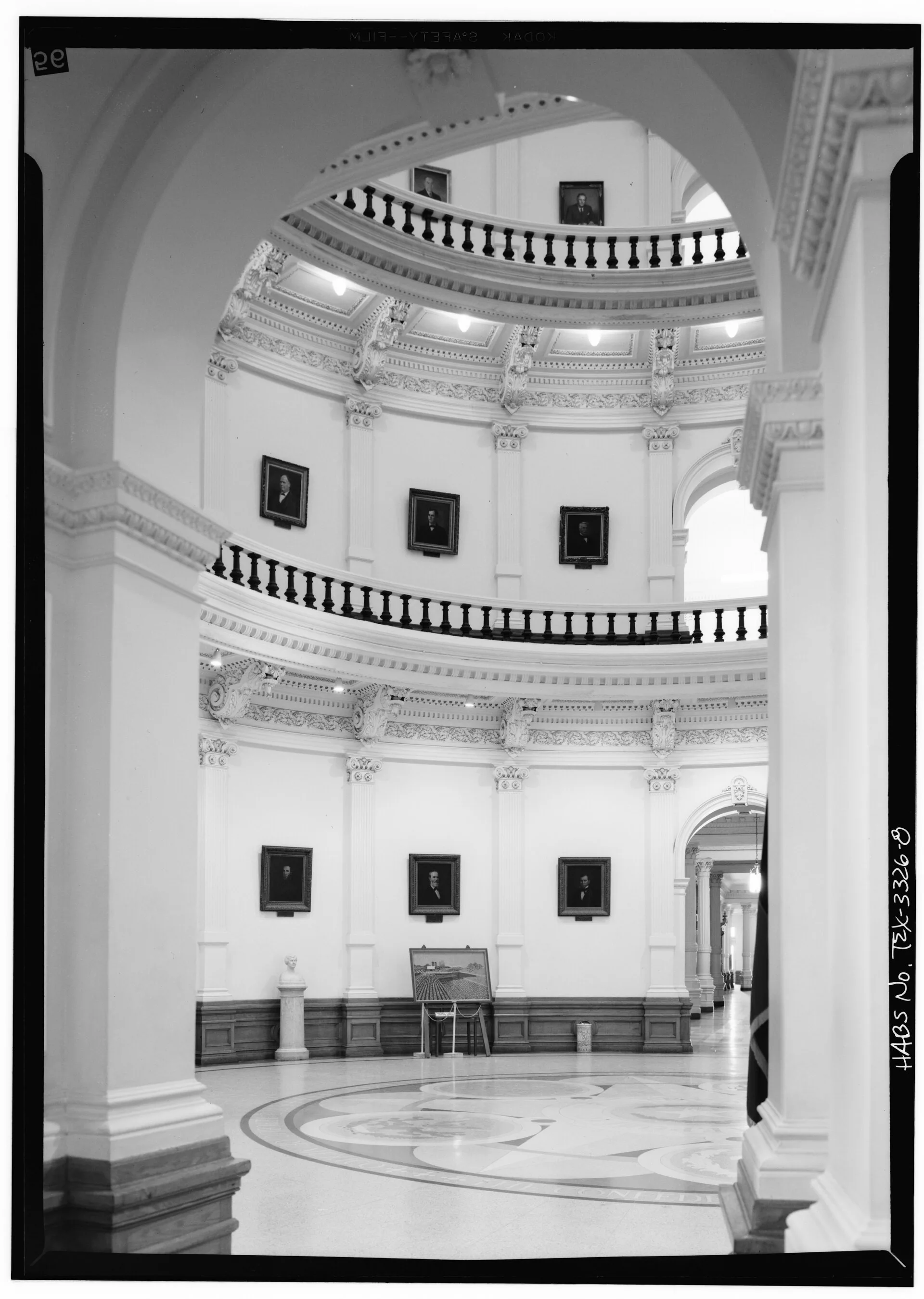 Black-and-white photograph of the Texas Capitol rotunda, showing tiers of balconies with railings and framed portraits rising above a patterned floor.