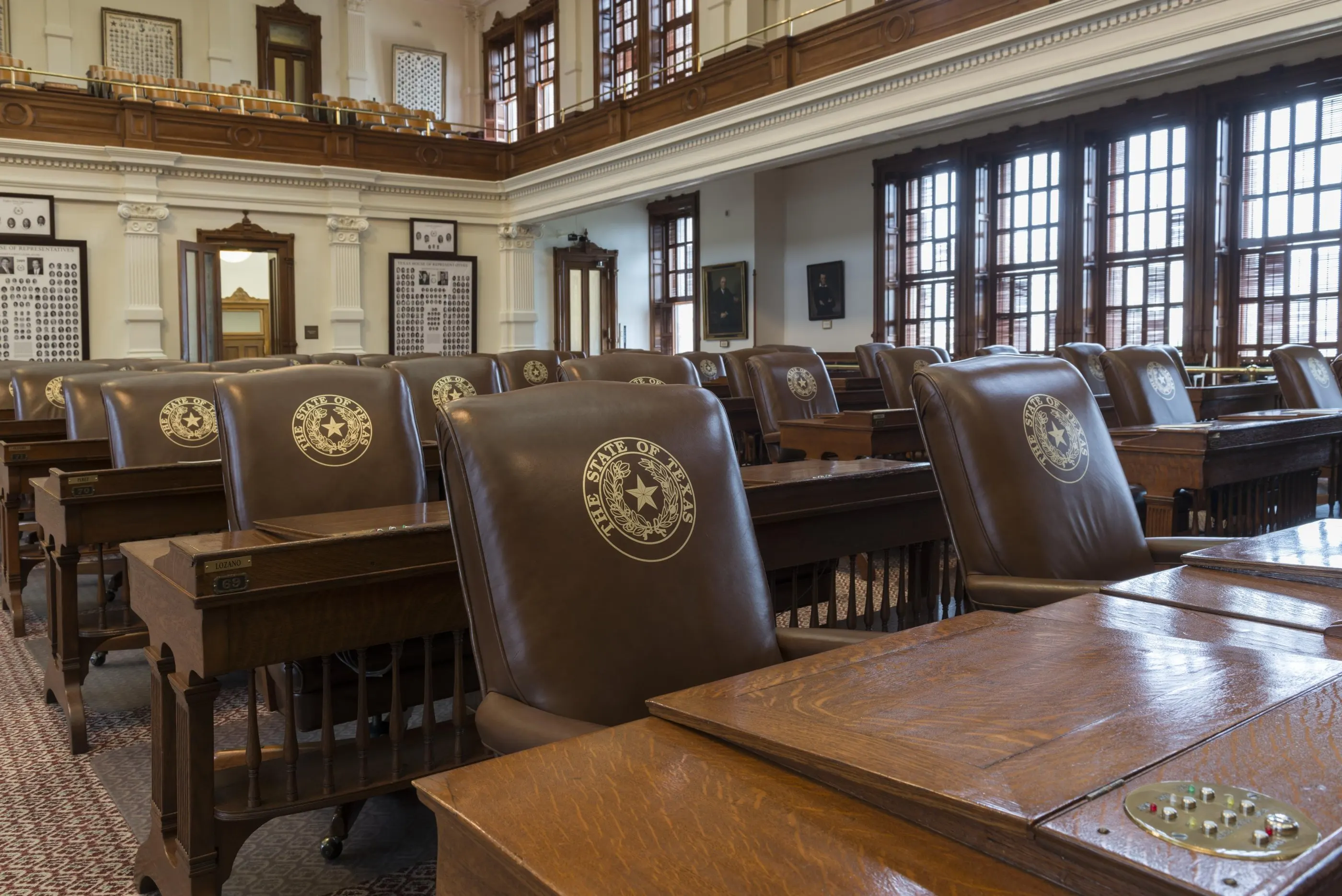 Rows of wooden desks and leather chairs bearing the seal of the State of Texas in the Texas House chamber, under tall windows.