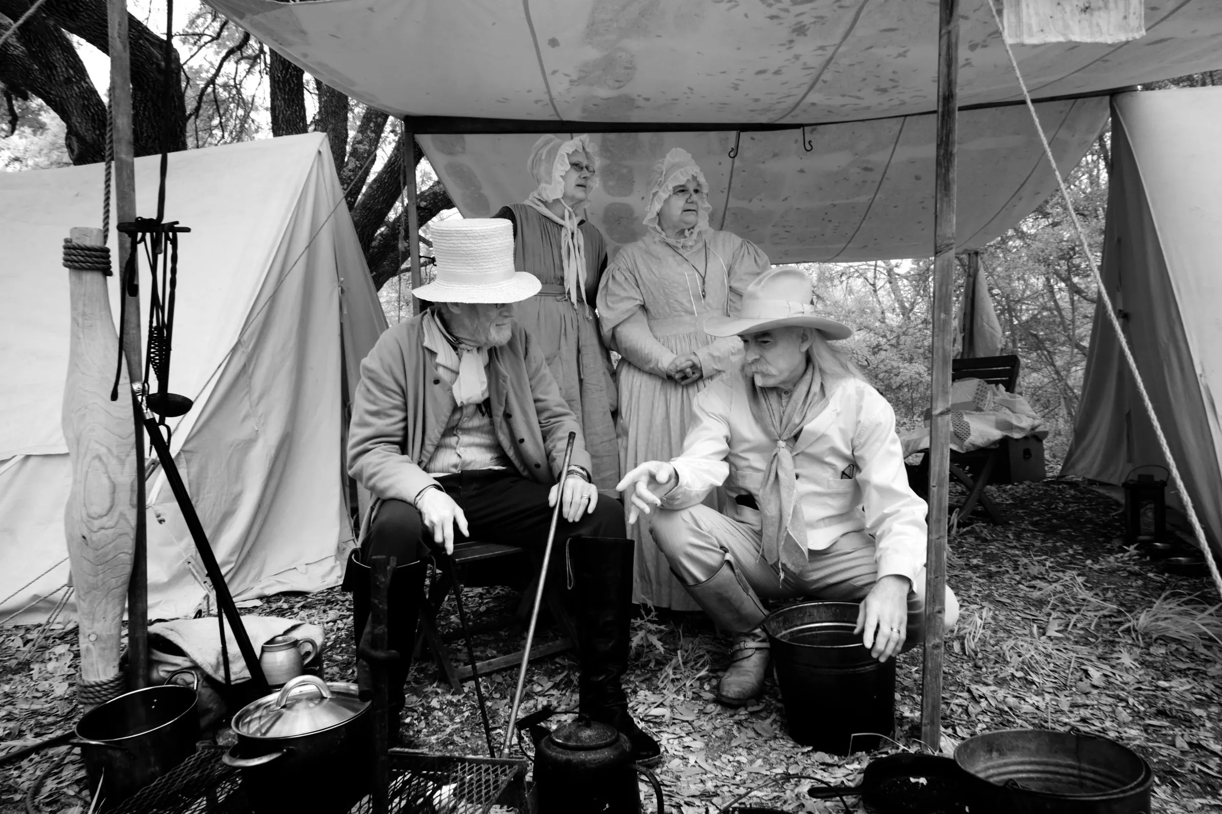 Black-and-white photograph of reenactors in 1830s dress seated under a canvas tent with cooking pots, at the site where the Texas Declaration of Independence was adopted.